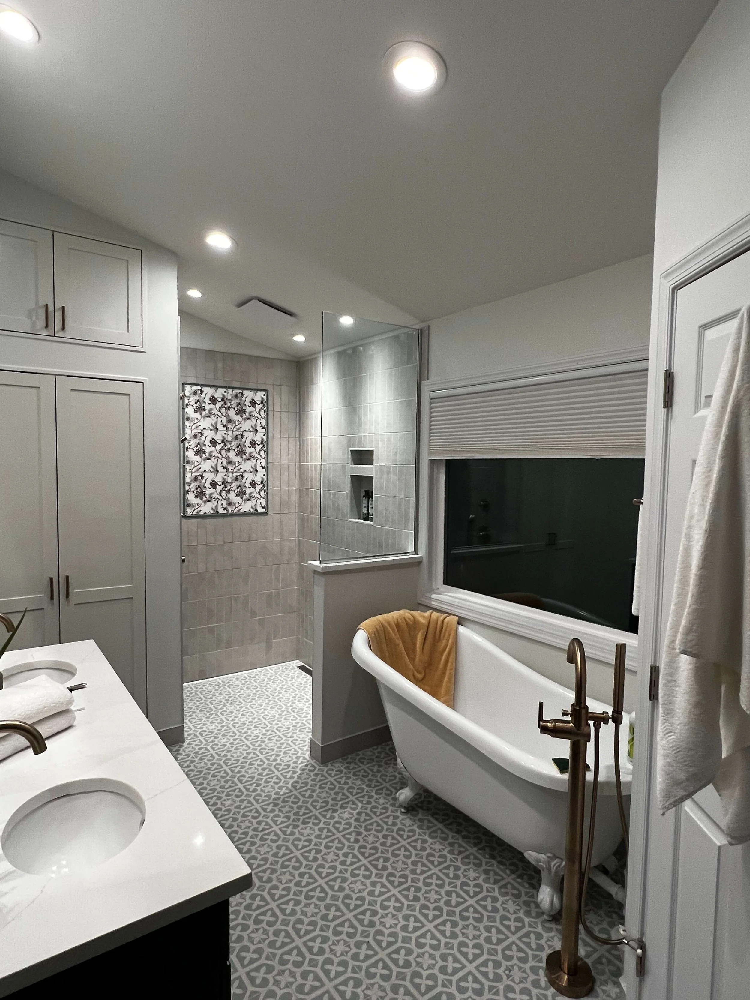 A bathroom featuring a clawfoot bathtub, a large window with a rolled-up shade, a shower with tiled walls separated by a glass partition, a white vanity with a marble countertop, and patterned floor tiles.