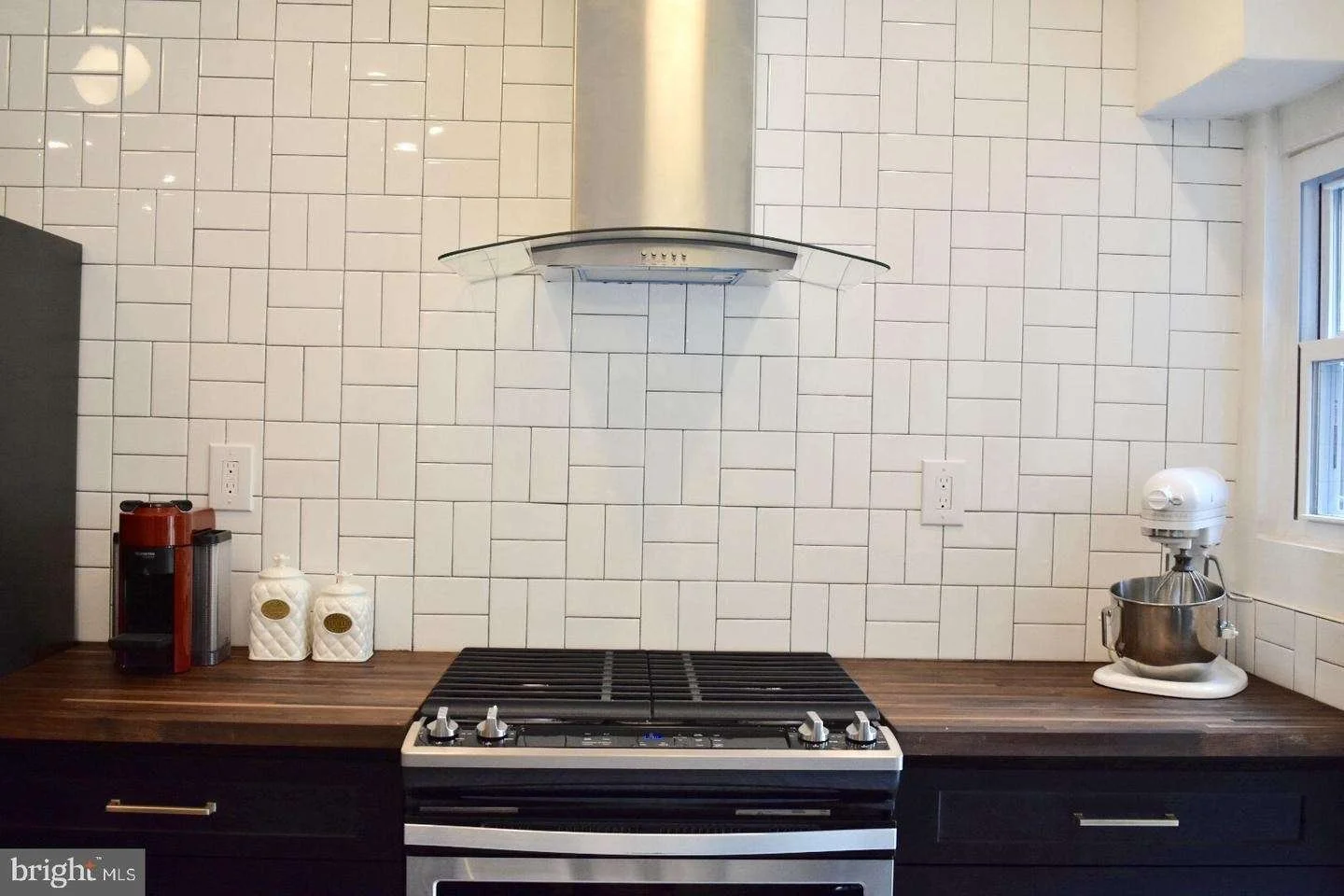 Kitchen with white subway tile backsplash, black cabinets, wooden countertop, stainless steel stove, range hood, coffee maker, white ceramic containers, and stand mixer near window.