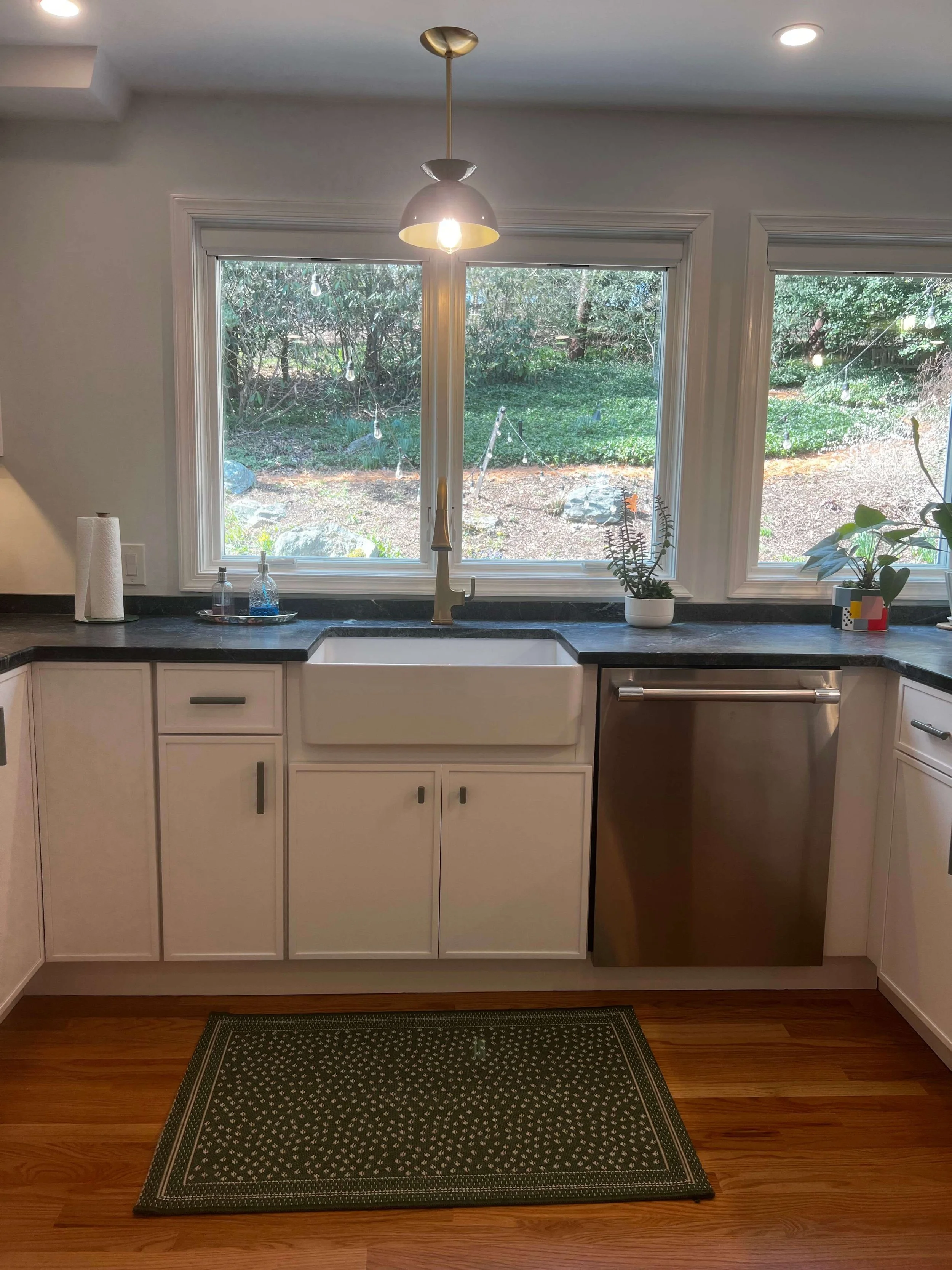 Kitchen with white cabinets, black countertop, white farmhouse sink, and a stainless steel dishwasher. Two large windows overlooking a garden, with a modern hanging light fixture and indoor plants on the countertop. Wooden floor and a green patterned