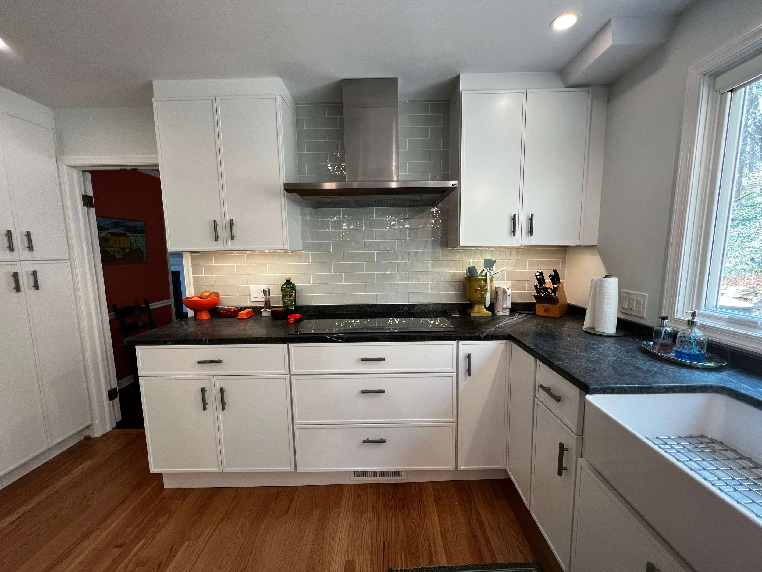 Kitchen with white cabinets, black countertops, gray tiled backsplash, stainless steel range hood, and a large window overlooking trees.