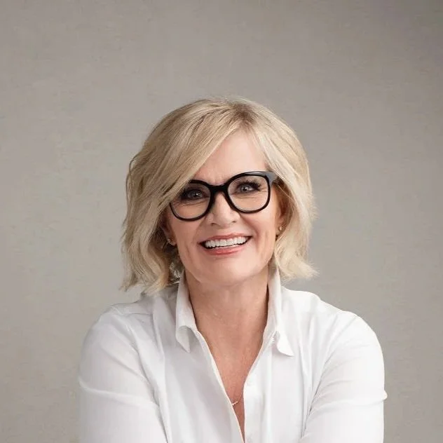 Portrait of a smiling middle-aged woman with blonde hair, black glasses, and a white shirt against a plain background.