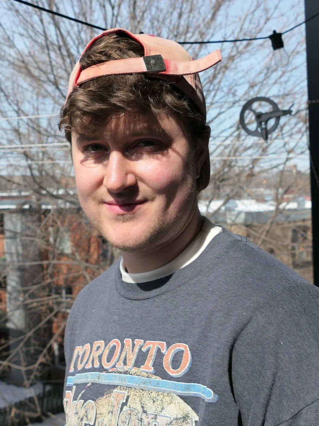 A young man with light brown hair, wearing a grey sweatshirt with a graphic print, a white undershirt, and a pink and beige baseball cap worn backwards. He is outdoors on a clear, sunny day with bare trees and houses in the background.