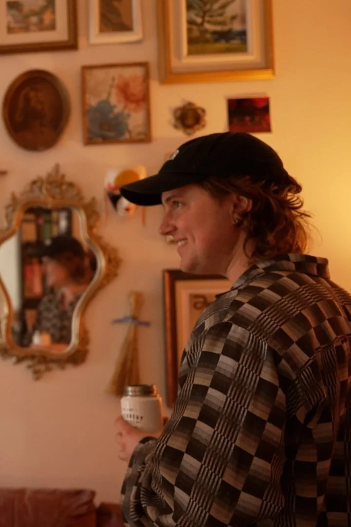Young man smiling in a patterned shirt and black cap holding a drink in a room decorated with various framed pictures and decorative items.
