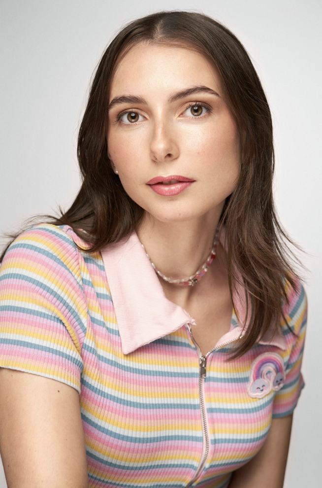 Young woman with brown hair wearing a colorful striped shirt with a rainbow patch, looking at the camera, against a plain light background.