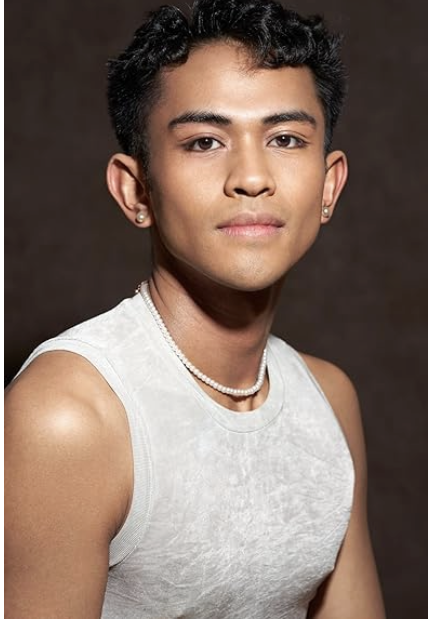 Portrait of a young person with dark, curly hair, earrings, a pearl necklace, and a sleeveless light-colored top against a dark background.