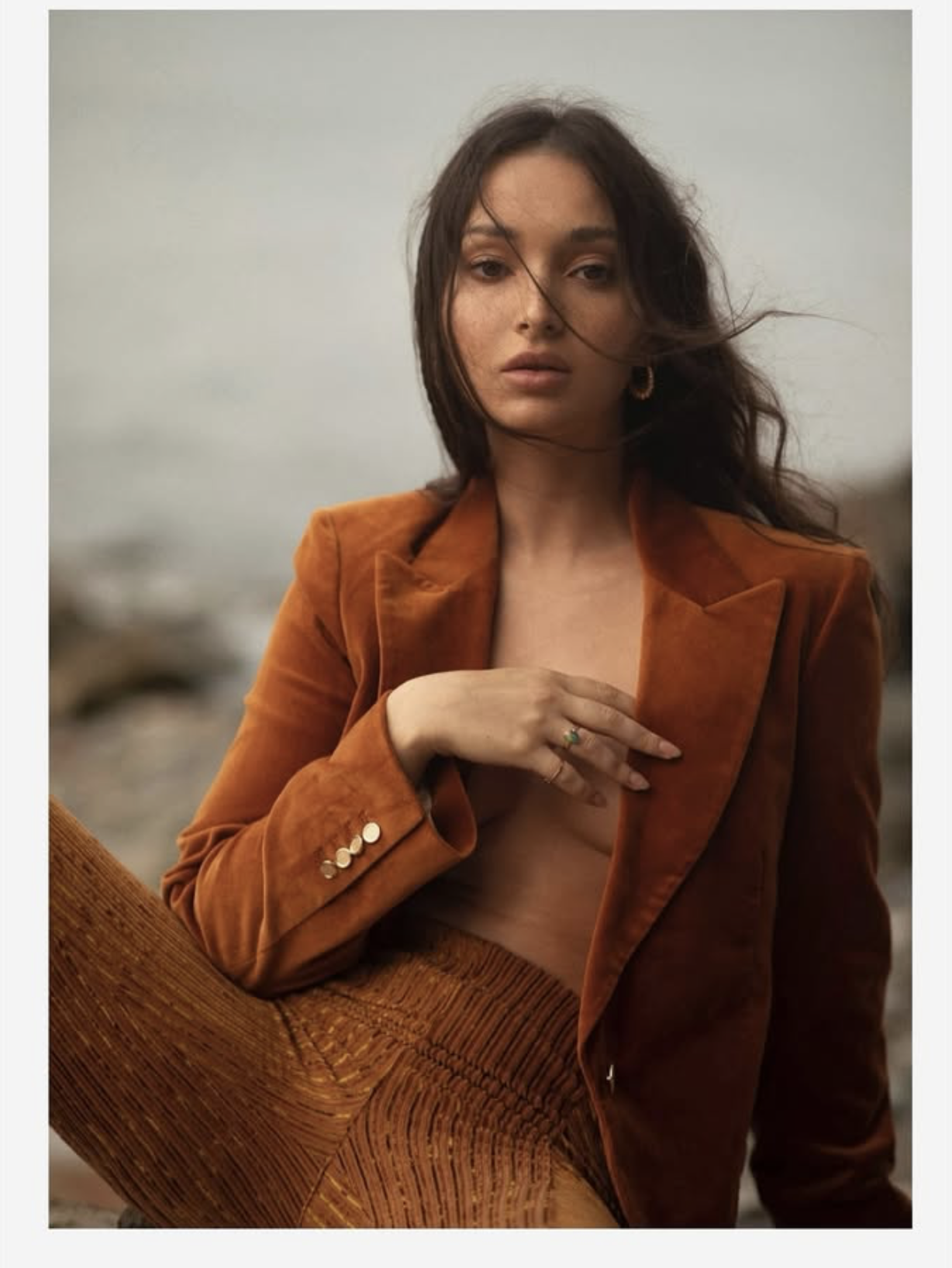 A woman with long brown hair and hoop earrings photographed outdoors with a cloudy sky and rocky landscape in the background. She is wearing an orange-brown blazer with gold buttons and matching textured pants, and is topless underneath the blazer. She has a calm, serious expression, and her hair is slightly tousled.