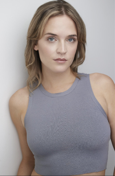 Young woman with shoulder-length wavy hair wearing a sleeveless gray top standing against a plain background