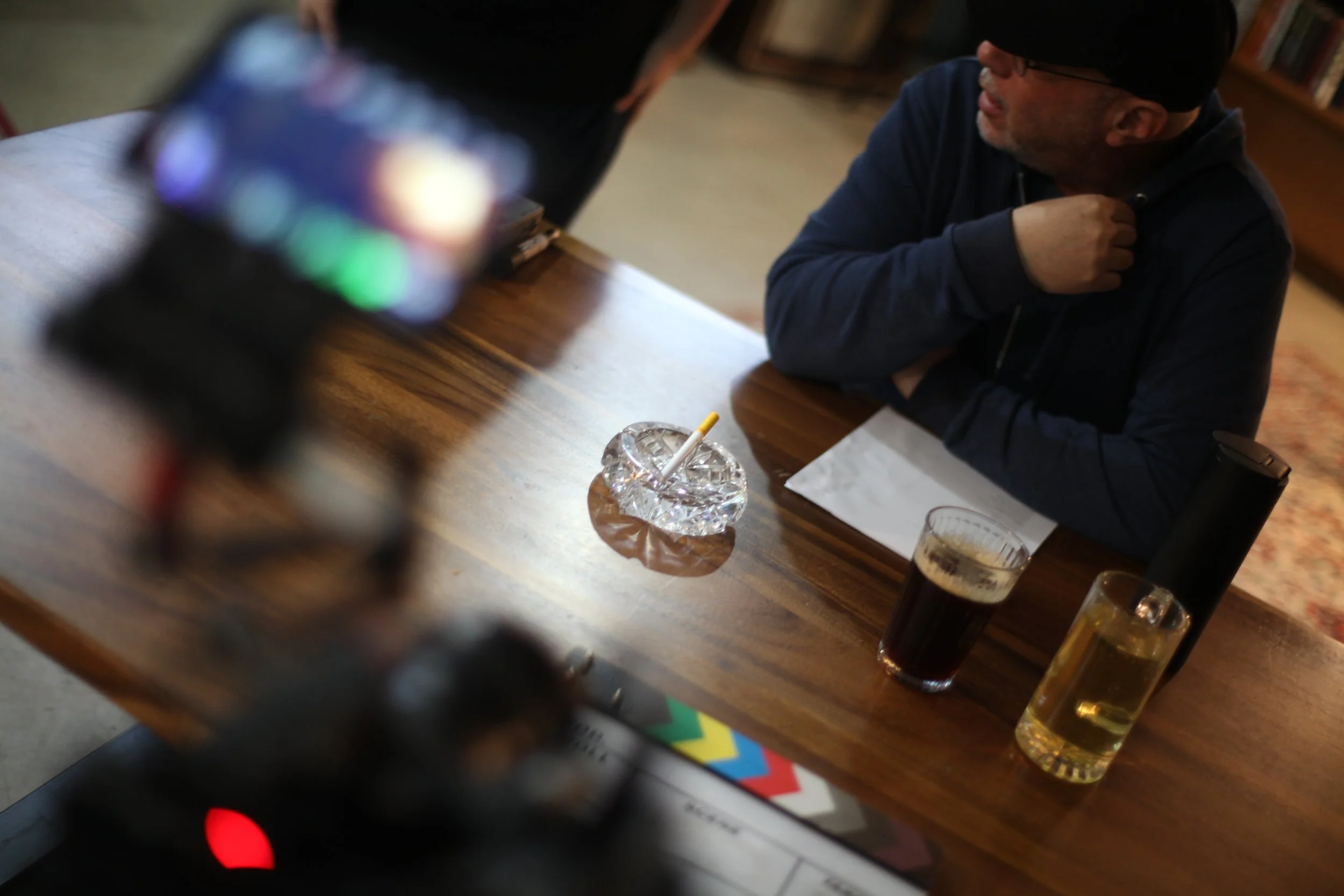 A man sitting at a wooden table with two drinks, one dark and one clear, an ashtray with a cigarette, a napkin, and a black speaker, with a blurred smartphone in the foreground, in a cozy room with a bookshelf.