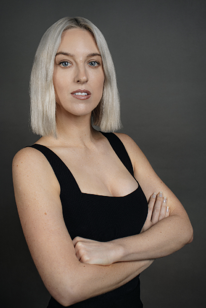 A woman with platinum blonde hair and blue eyes wearing a black sleeveless top with her arms crossed, posed against a gray background.