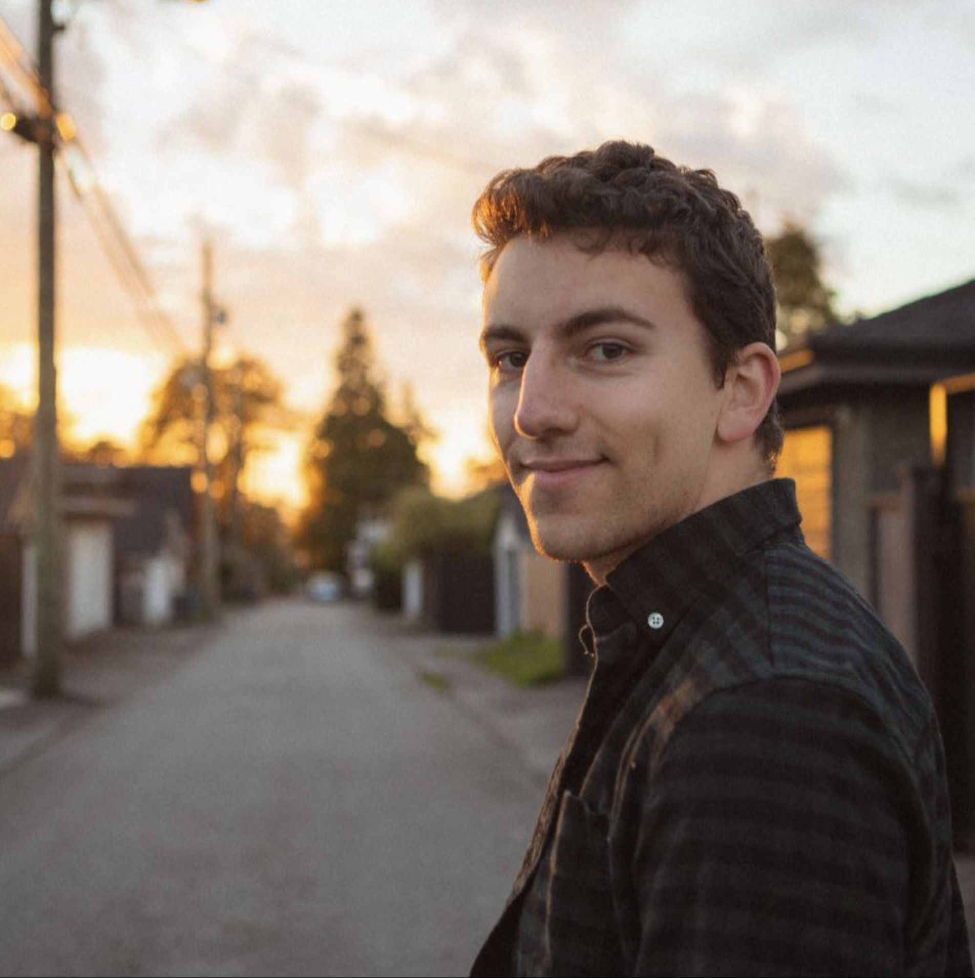 A young man with short, dark curly hair and light skin, smiling slightly, standing on an empty residential street at sunset. The background features houses, utility poles, and a cloudy sky illuminated by the setting sun.