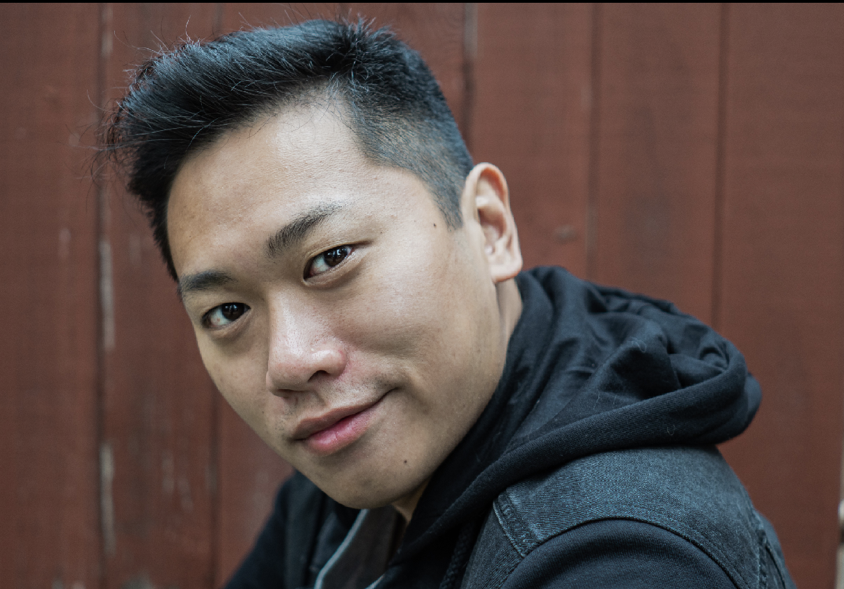 Close-up of a young Asian man with short black hair smiling, wearing a black hoodie, against a wooden background.
