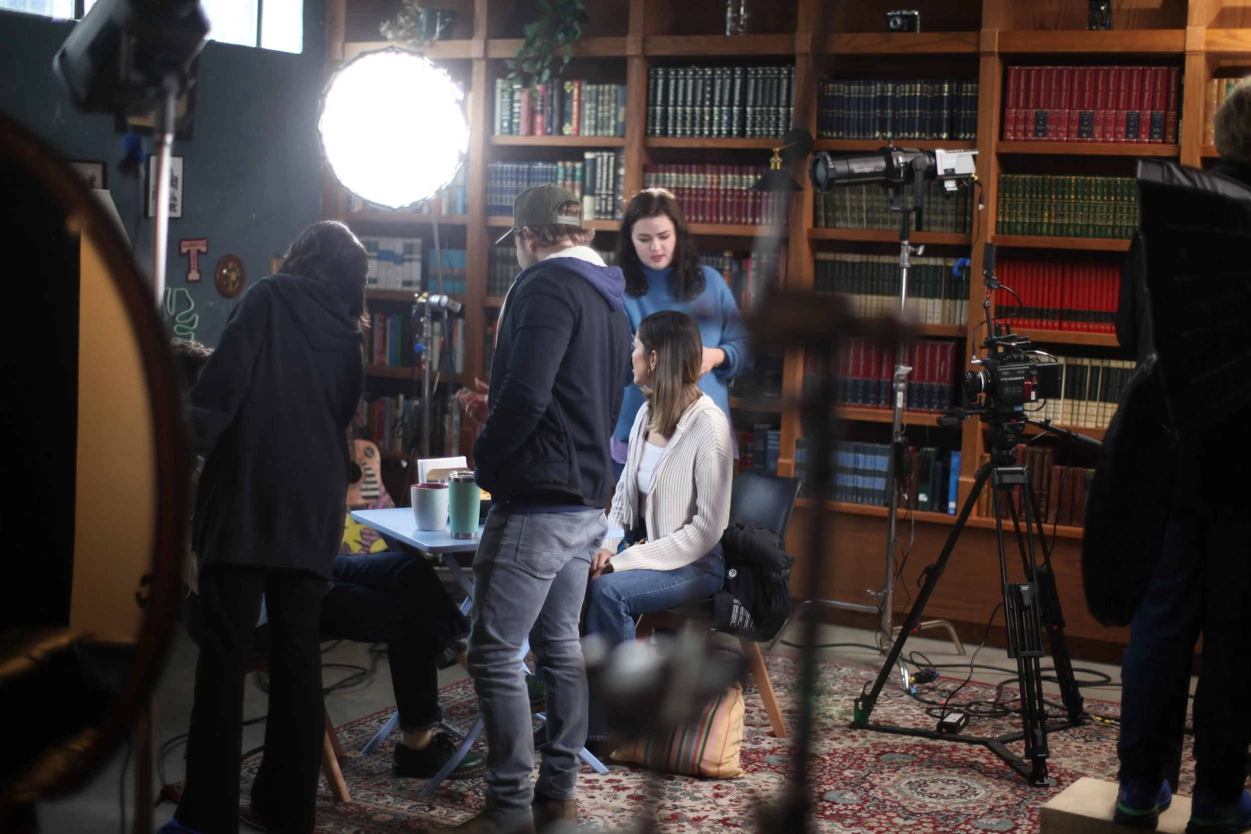 A film crew setting up for a shoot in a cozy library or study room with wooden bookshelves filled with books. Crew members are gathered around a woman sitting at a table with cups, while others prepare equipment and light the scene.