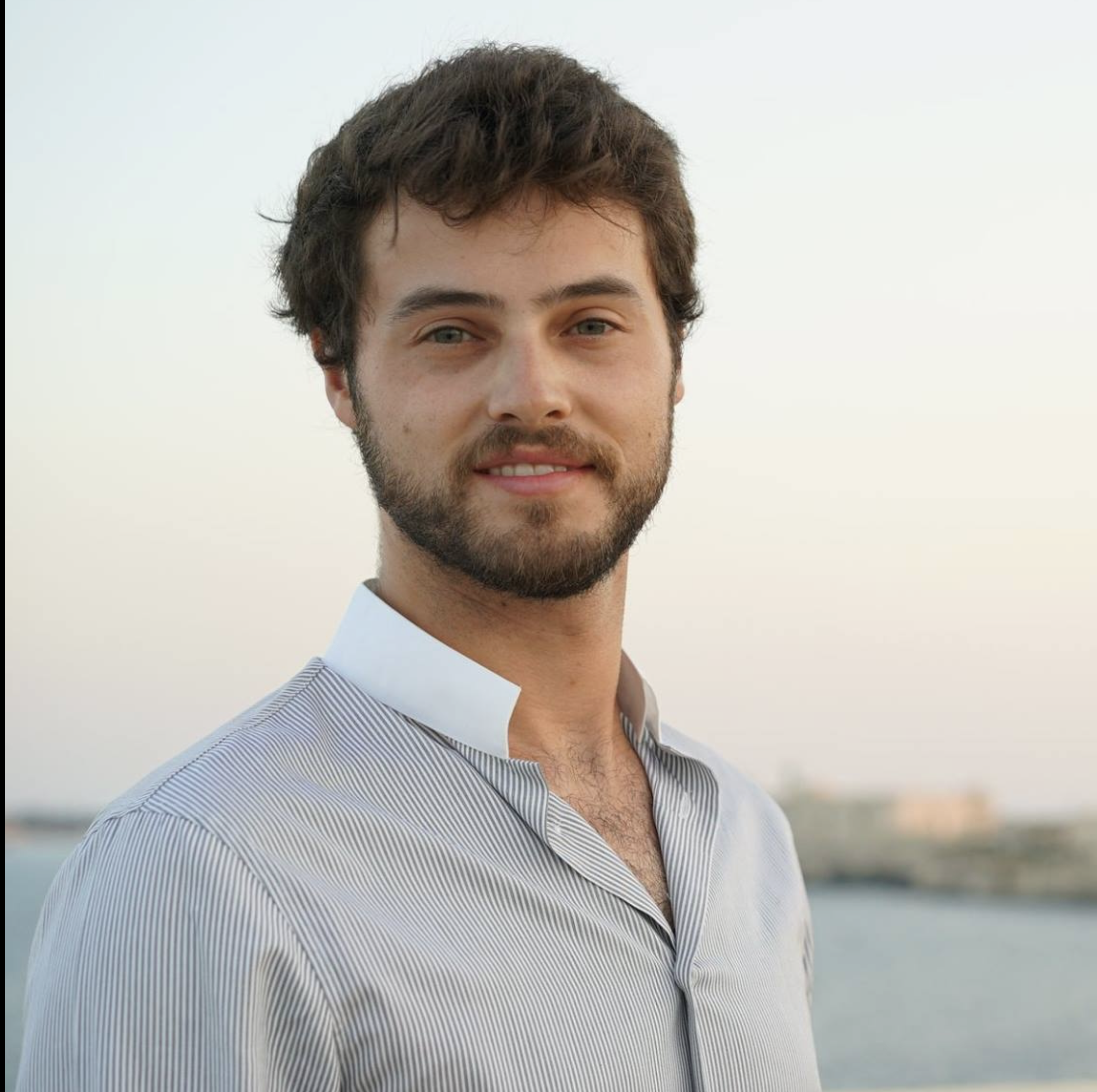 A young man with brown hair and a beard standing outdoors near water during sunset.