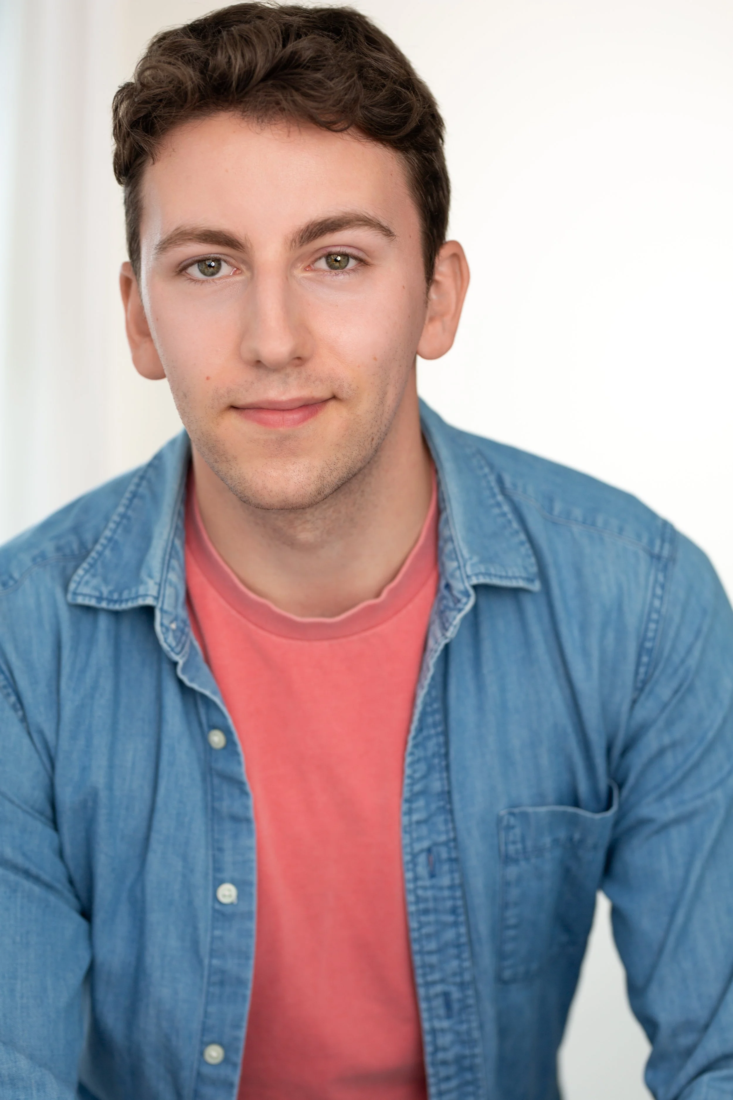Young man with short brown hair and light skin wearing a pink t-shirt and a blue denim button-up shirt, smiling softly against a plain light background.