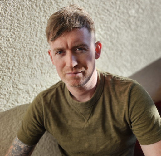A young man with light brown hair wearing a green T-shirt, sitting indoors against a beige textured wall, looking into the camera with a slight smile.