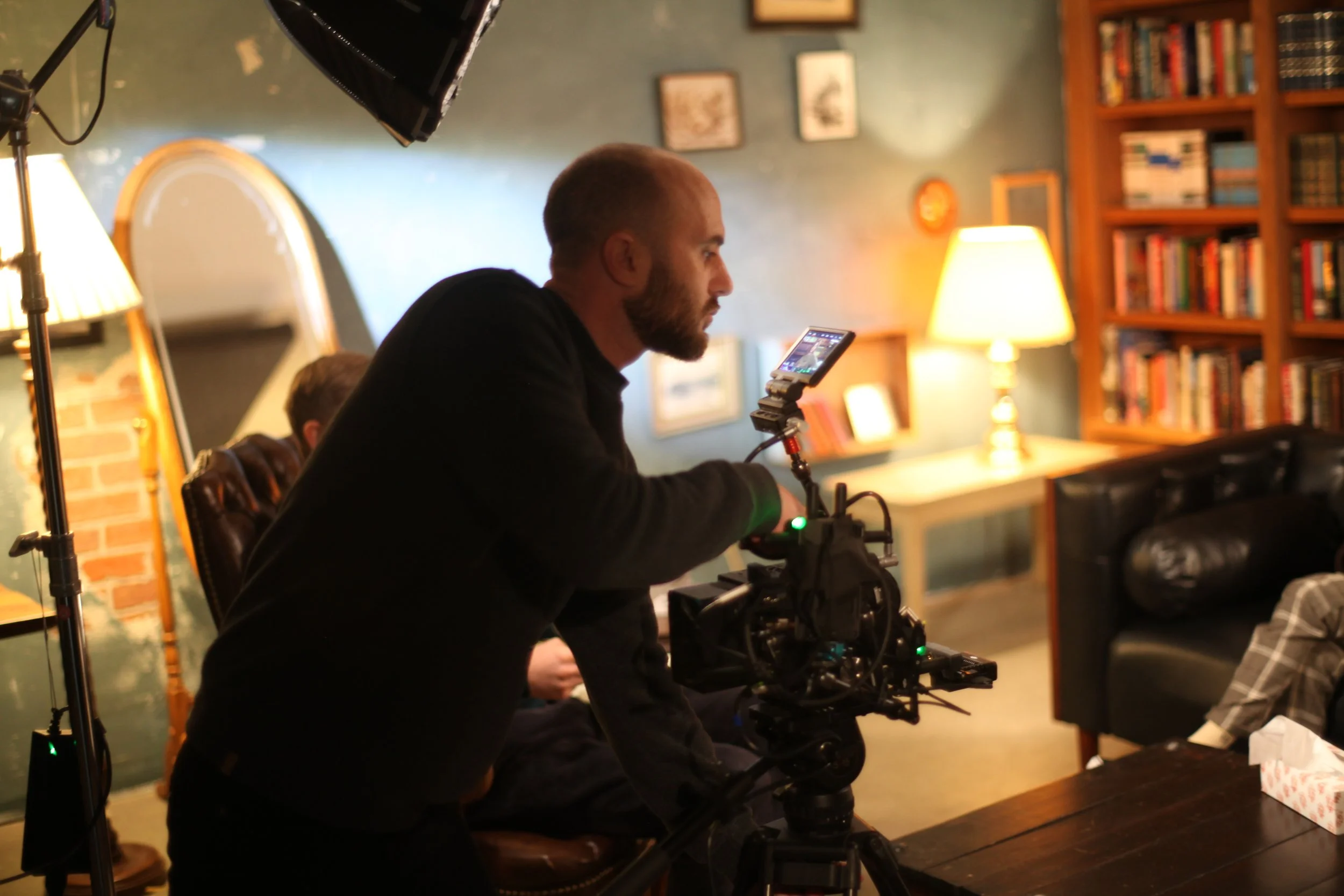 A man operating a professional film camera indoors with bookshelves, lamps, and artwork in the background.