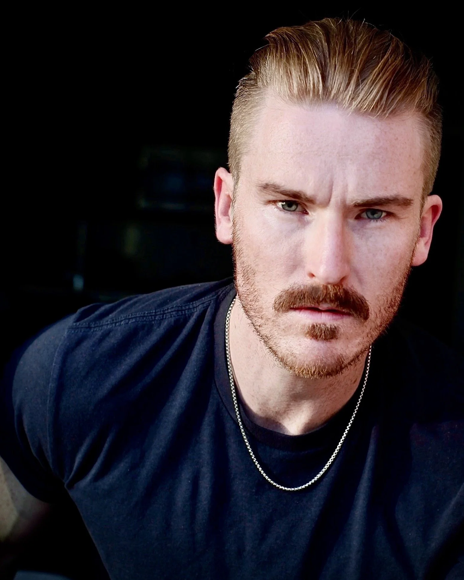 Close-up of a man with light skin, blond hair slicked back, a mustache, and beard, wearing a black shirt and a silver chain necklace, against a dark background.