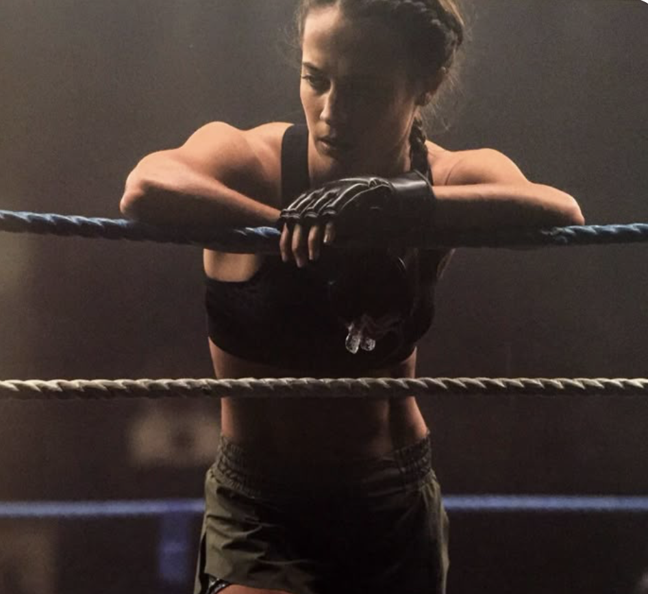 A female boxer leaning on the ropes of a boxing ring, wearing black sports attire and boxing gloves, against a dark background.
