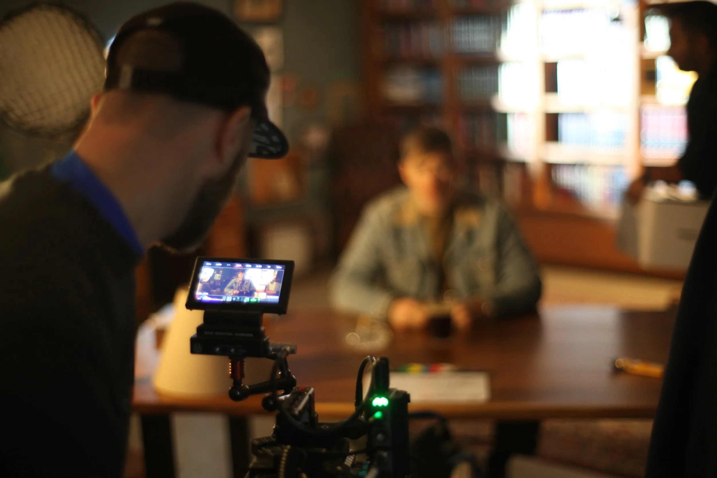 A person filming a woman sitting at a table in a room with bookshelves. The woman is slightly out of focus, and the filming person is in focus, with a camera on a stabilizer in the foreground.