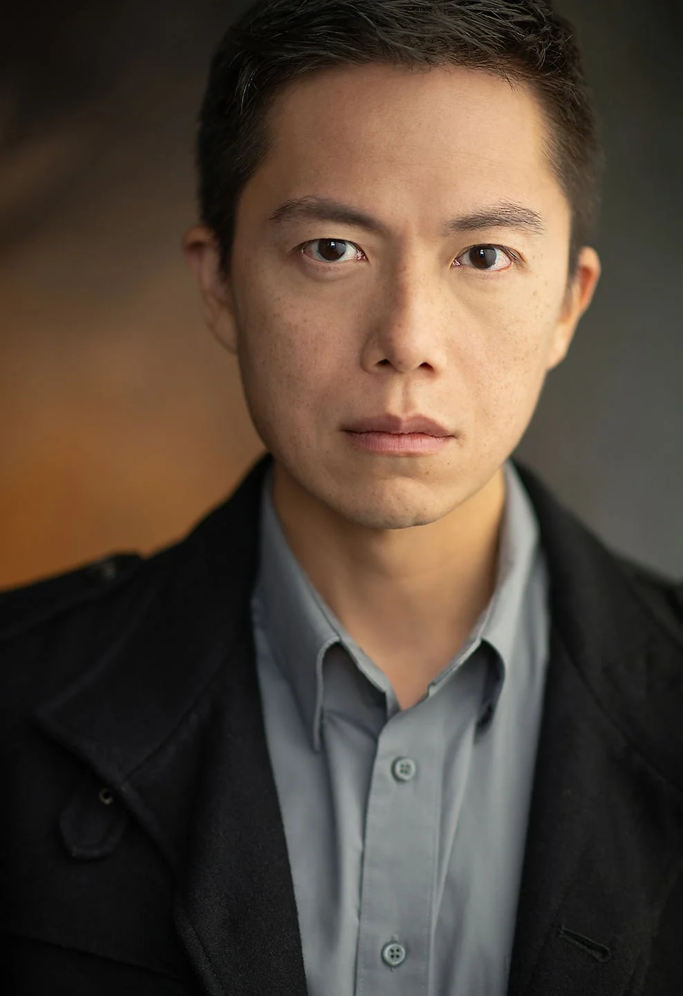 Close-up portrait of a young man with dark hair wearing a gray shirt and black jacket, looking serious, against a blurred dark background.
