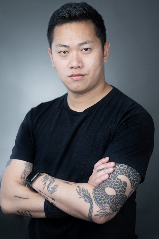 A young man with short black hair and tattoos on his arms, wearing a black T-shirt and a smartwatch, standing with arms crossed against a plain gray background.