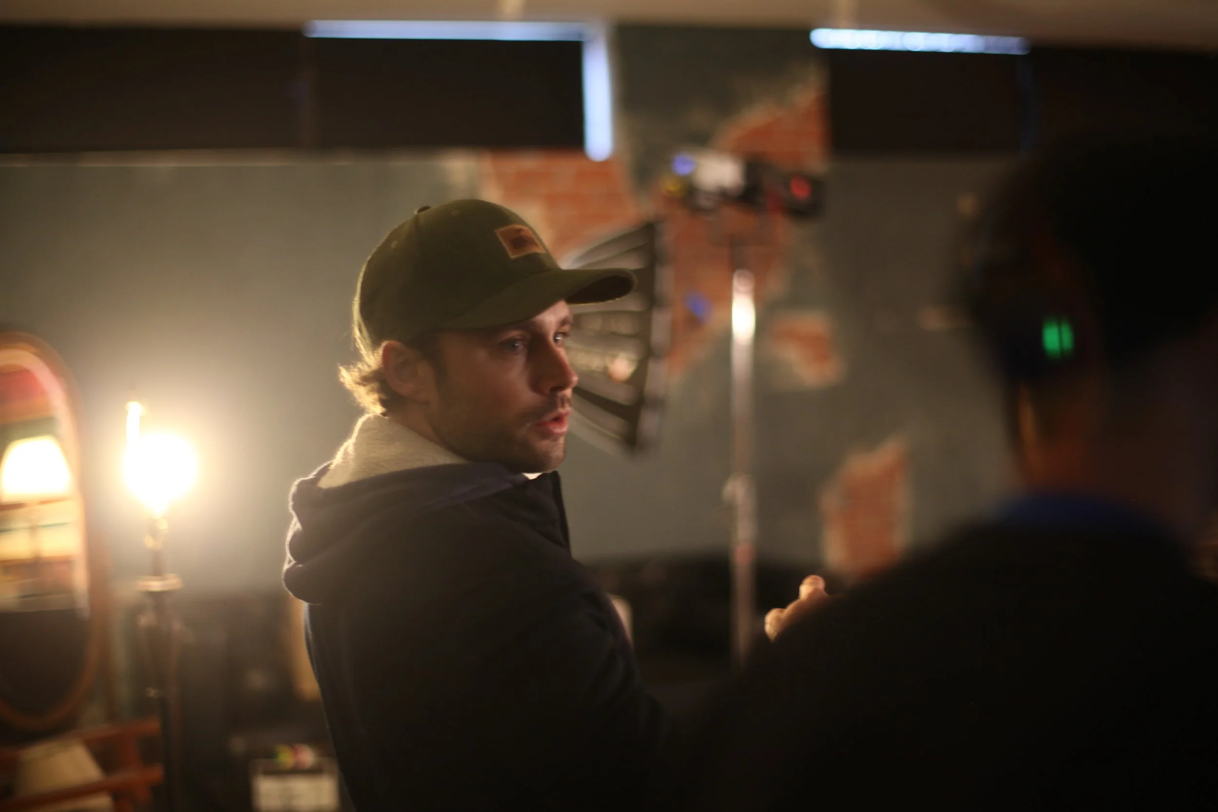 A man wearing a baseball cap and hoodie engaging in a conversation with another person on a film set, with professional lighting equipment in the background.