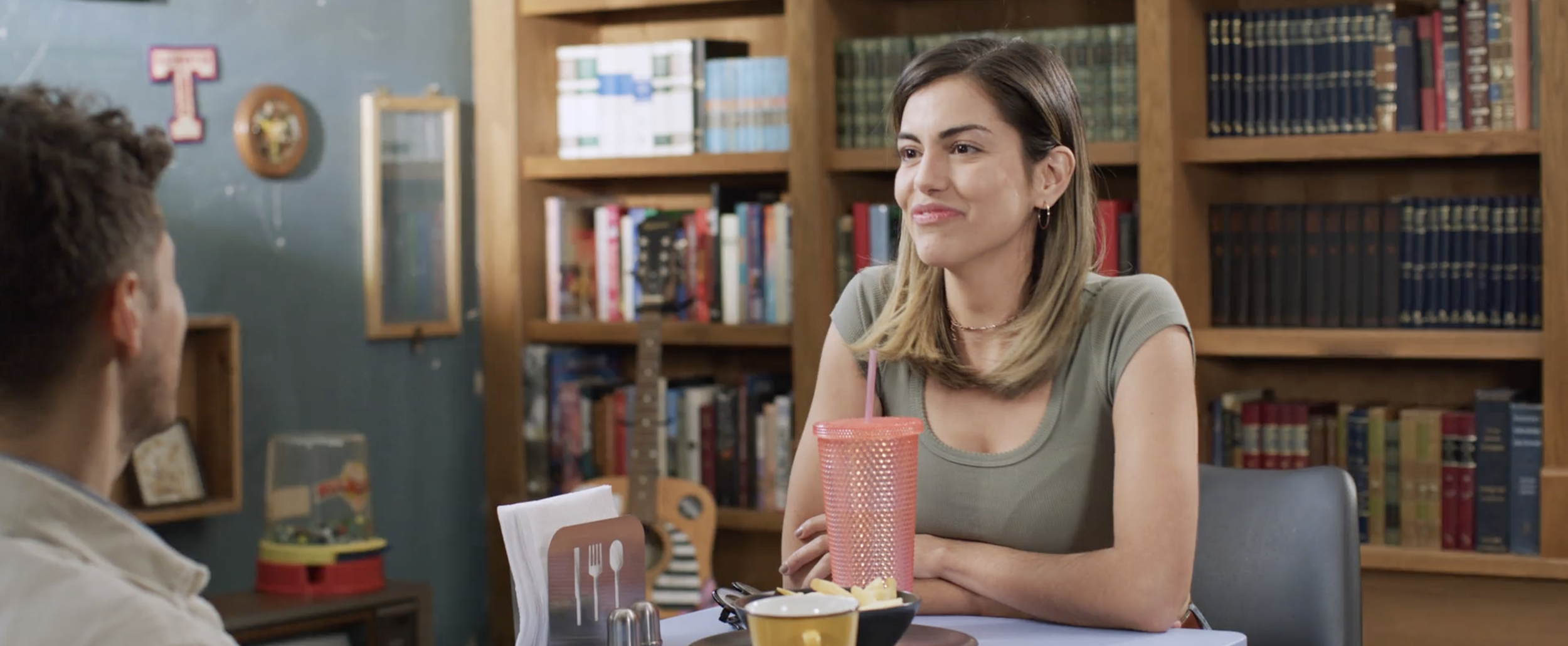 A woman with shoulder-length light brown hair and earrings sitting at a table in a cozy, book-filled room, smiling and looking at a man whose back is facing the camera. The table has a pink cup with a straw, a bowl of food, and a yellow cup.