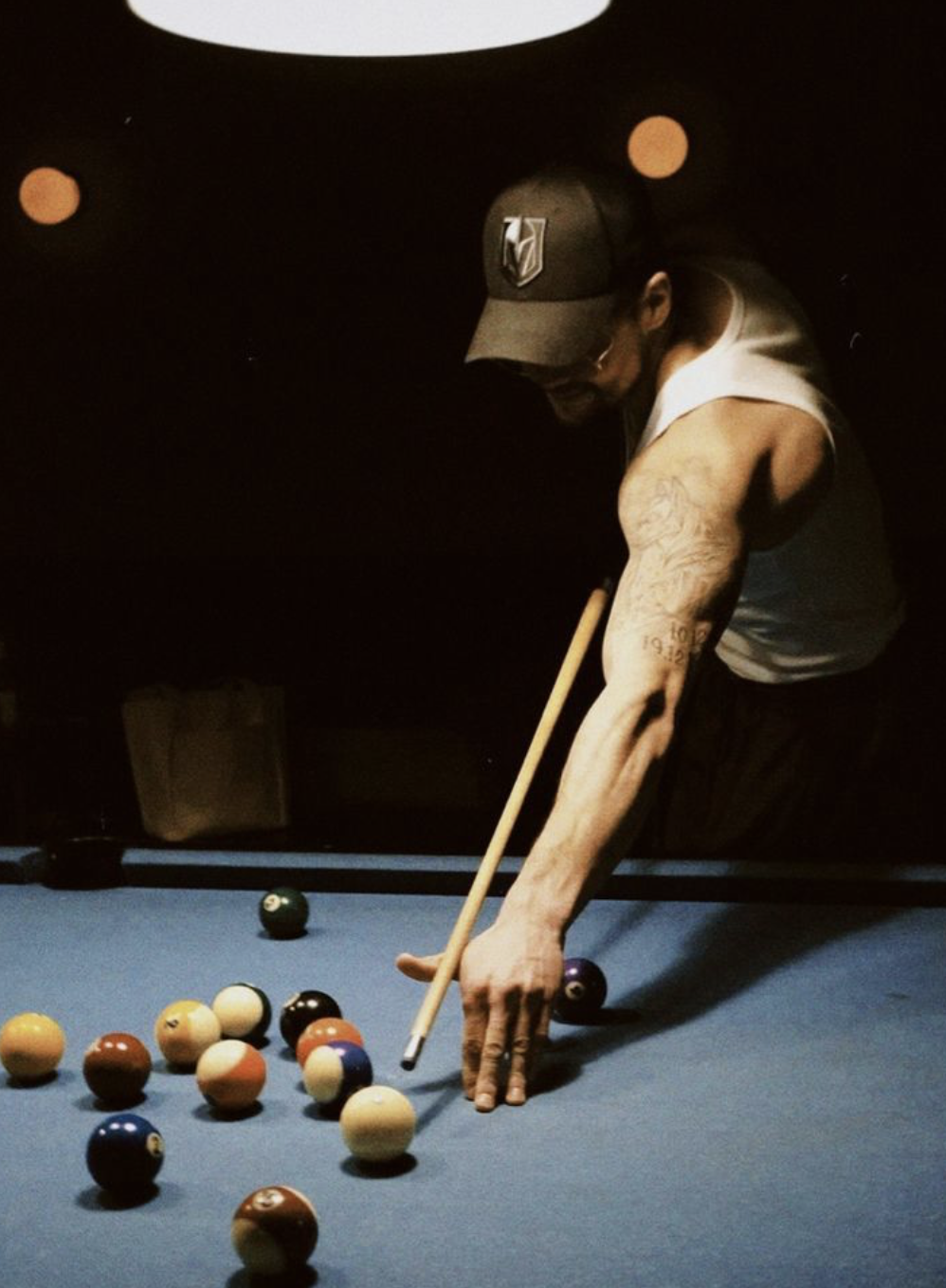 Person playing pool on a blue-felt billiards table, wearing a baseball cap and sleeveless shirt, aiming with a cue stick at the striped balls.