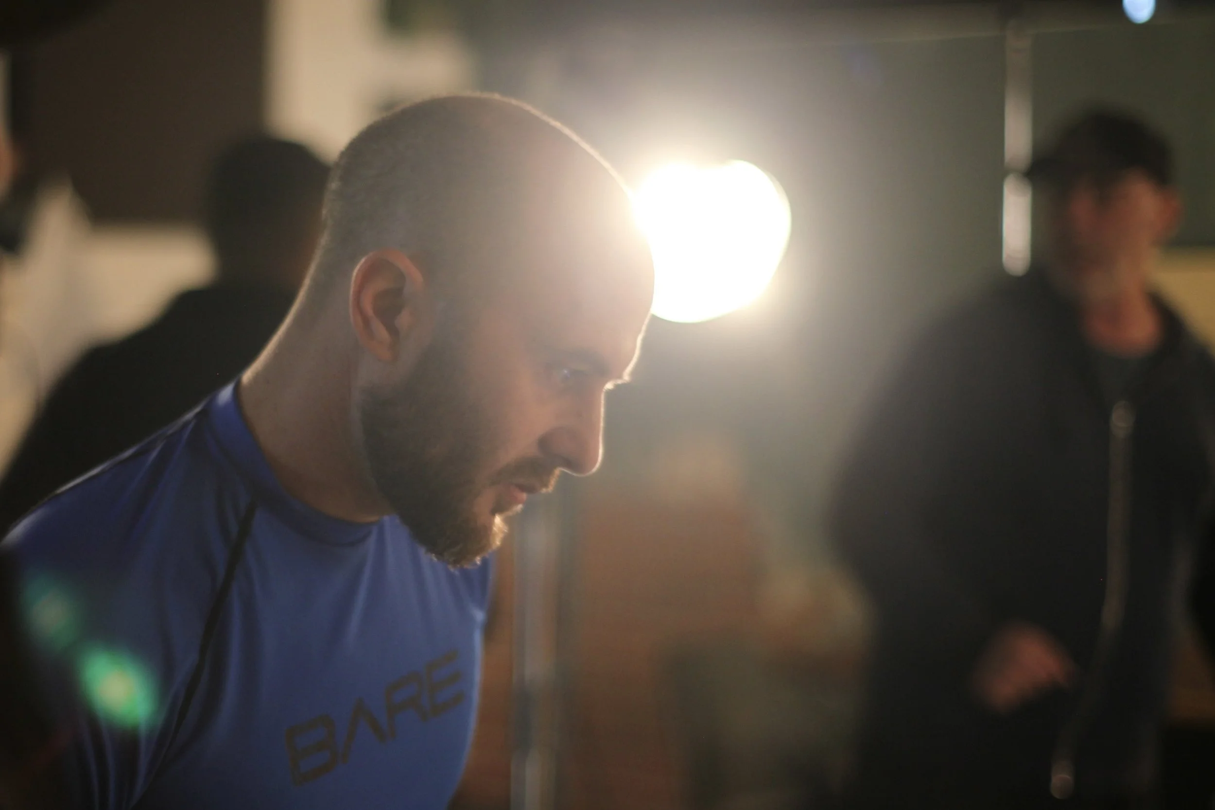 A man with a beard and a shaved head wearing a blue sports shirt, looking down thoughtfully in an indoor setting with warm lighting, and blurred background figures.