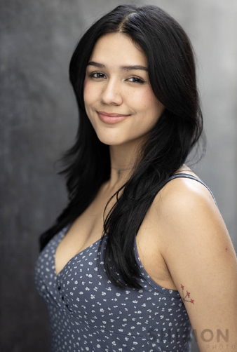 A young woman with long black hair, wearing a blue polka dot sleeveless top, smiling slightly, against a blurred gray background.