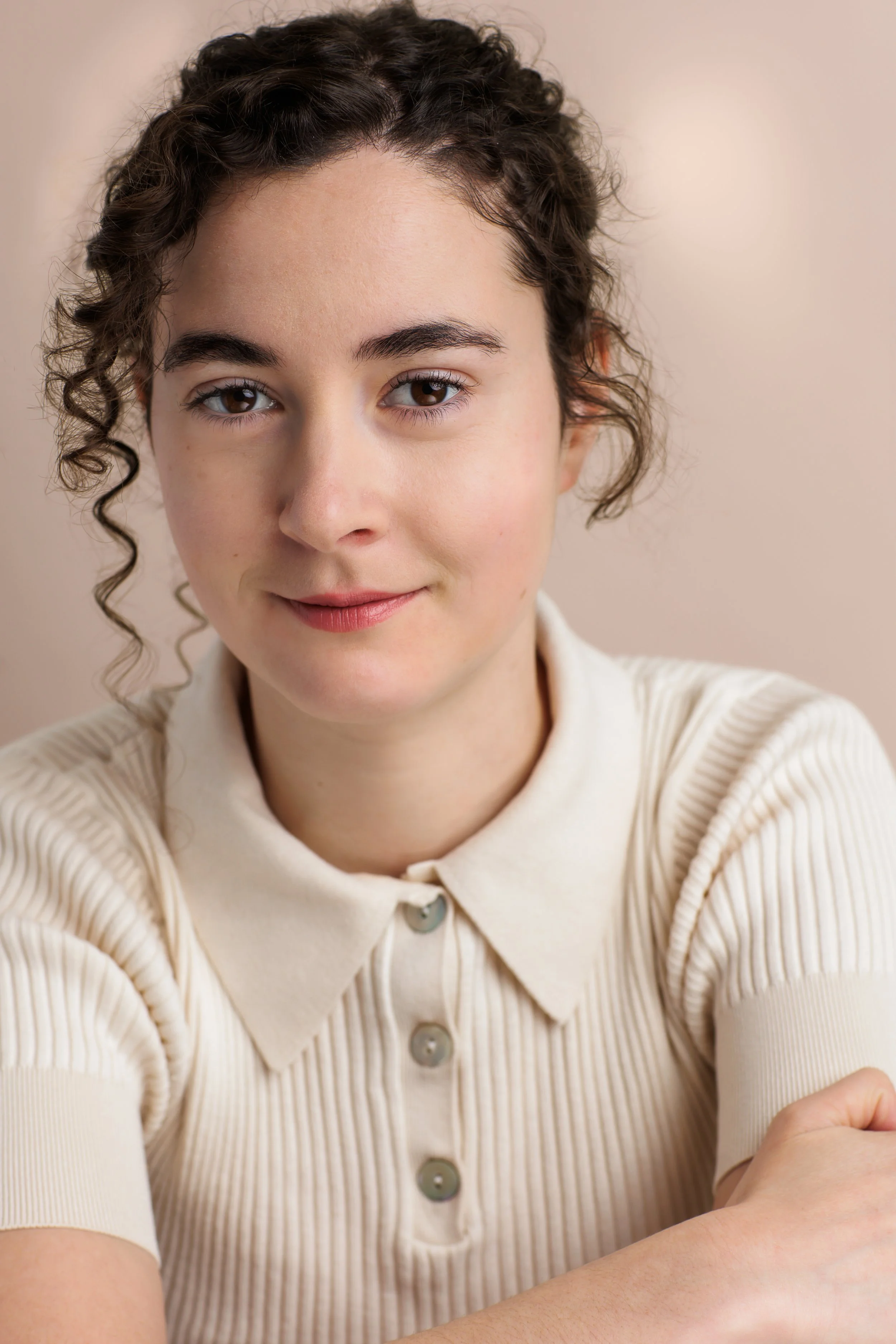 Close-up portrait of a young woman with curly brown hair, wearing a cream-colored, short-sleeved, collared shirt, smiling softly at the camera.
