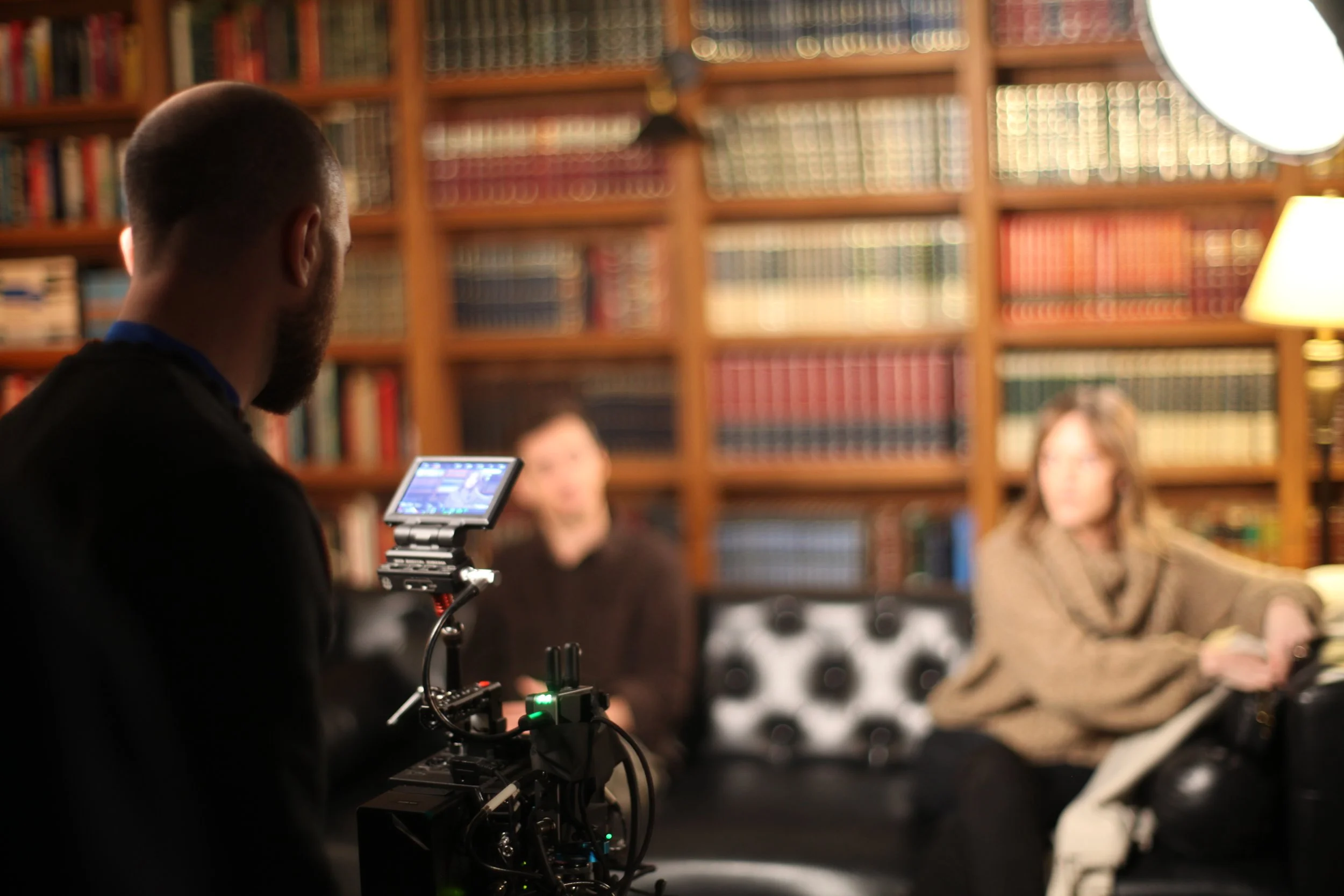 A film director operating a camera on a tripod, filming two women sitting on a black leather sofa in a room with wooden bookshelves filled with books, with a lamp to the right.