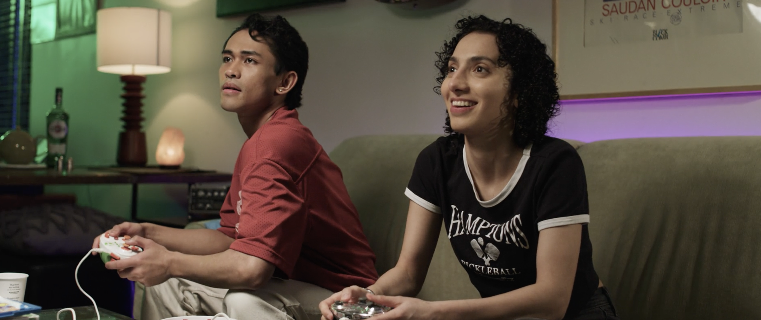 Two young women sitting on a couch playing video games, one holding a gaming controller and the other a game controller, in a cozy room with soft lighting, a lamp, and decorative items in the background.