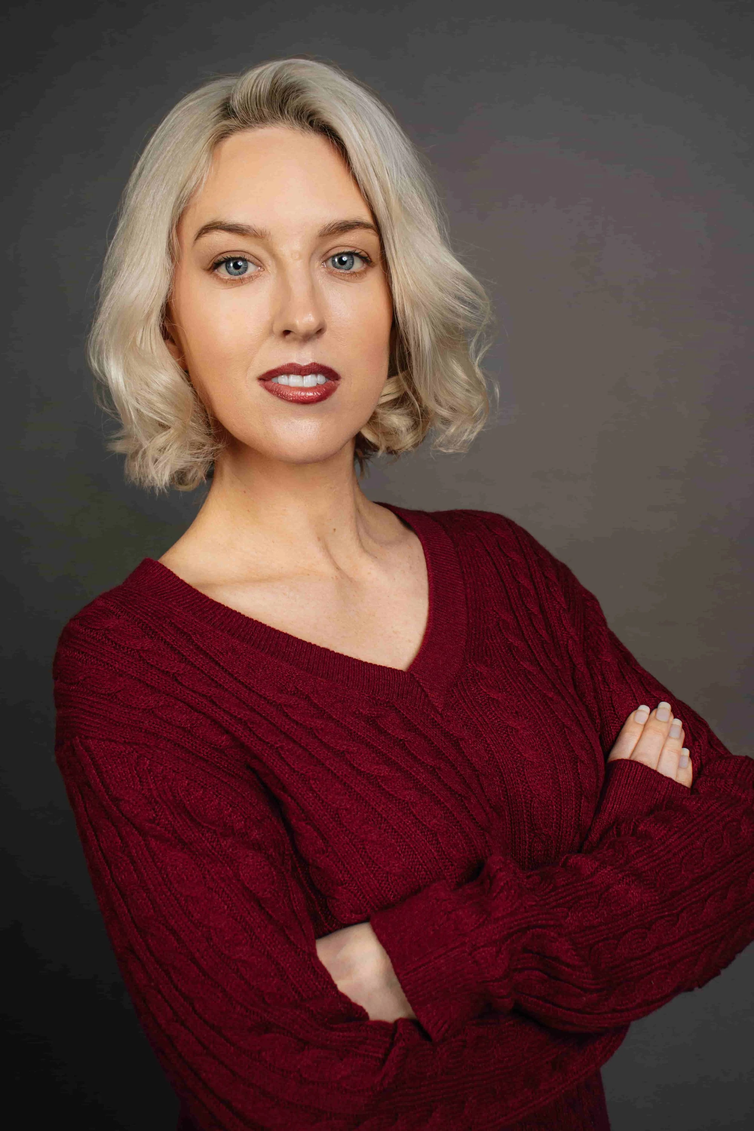 A woman with blonde hair and blue eyes wearing a red sweater with crossed arms looking at the camera, standing against a dark gray background.