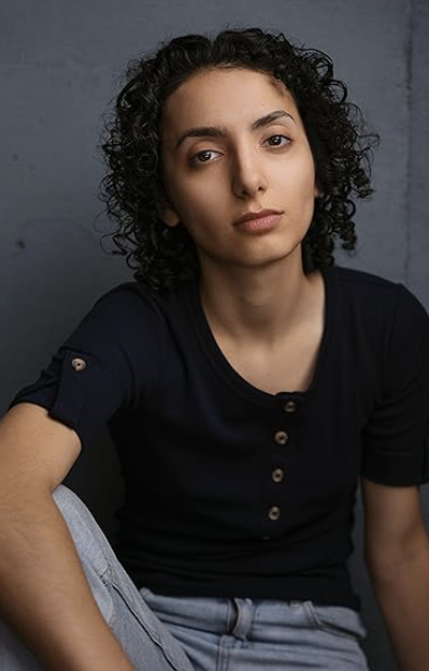 Portrait of a young woman with curly dark hair wearing a black top, sitting against a gray background.