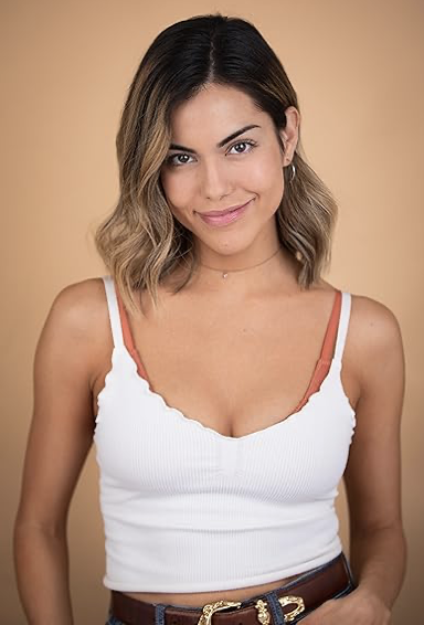 A young woman with shoulder-length wavy hair wearing a white tank top and a subtle smile, standing against a beige background.
