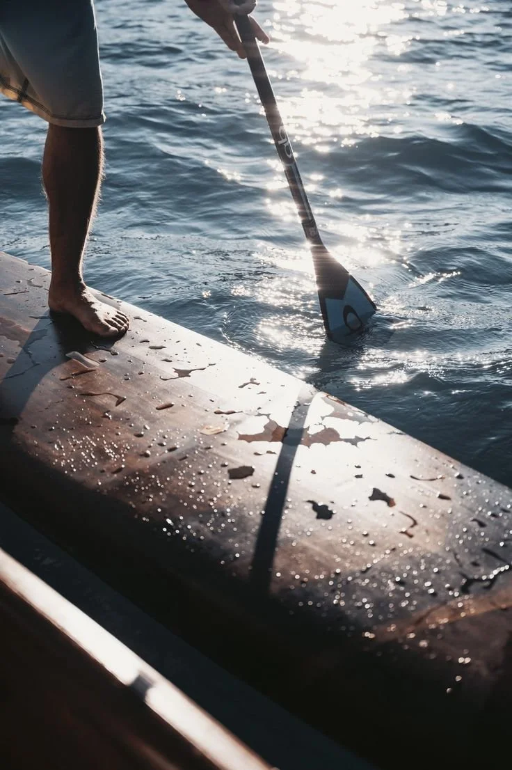 Person standing barefoot on dock, using paddleboard paddle to glide in water, sunlight reflecting off water surface.