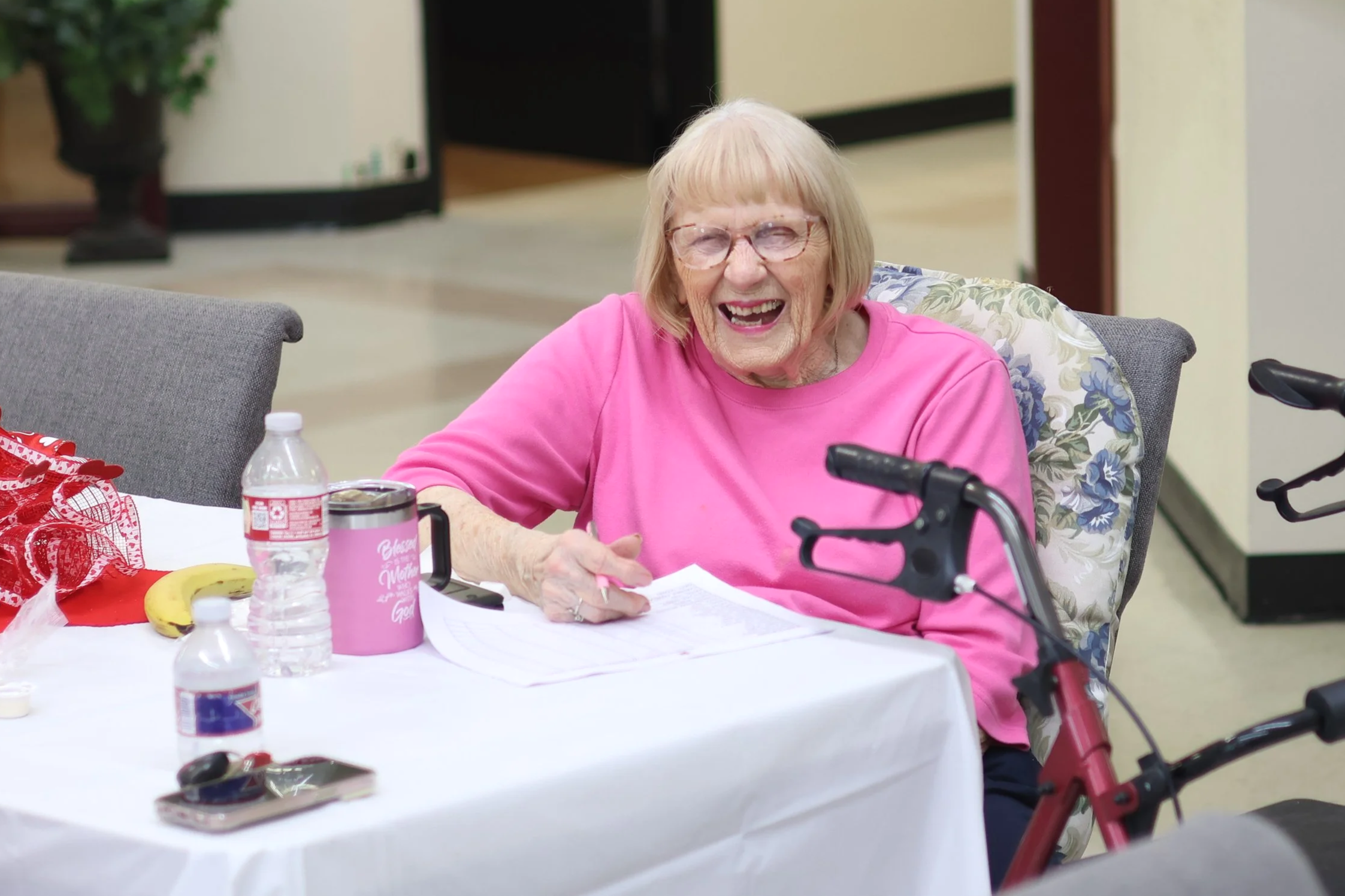 Old woman with glasses smiling, sitting at a table with a walker nearby, wearing a pink shirt.