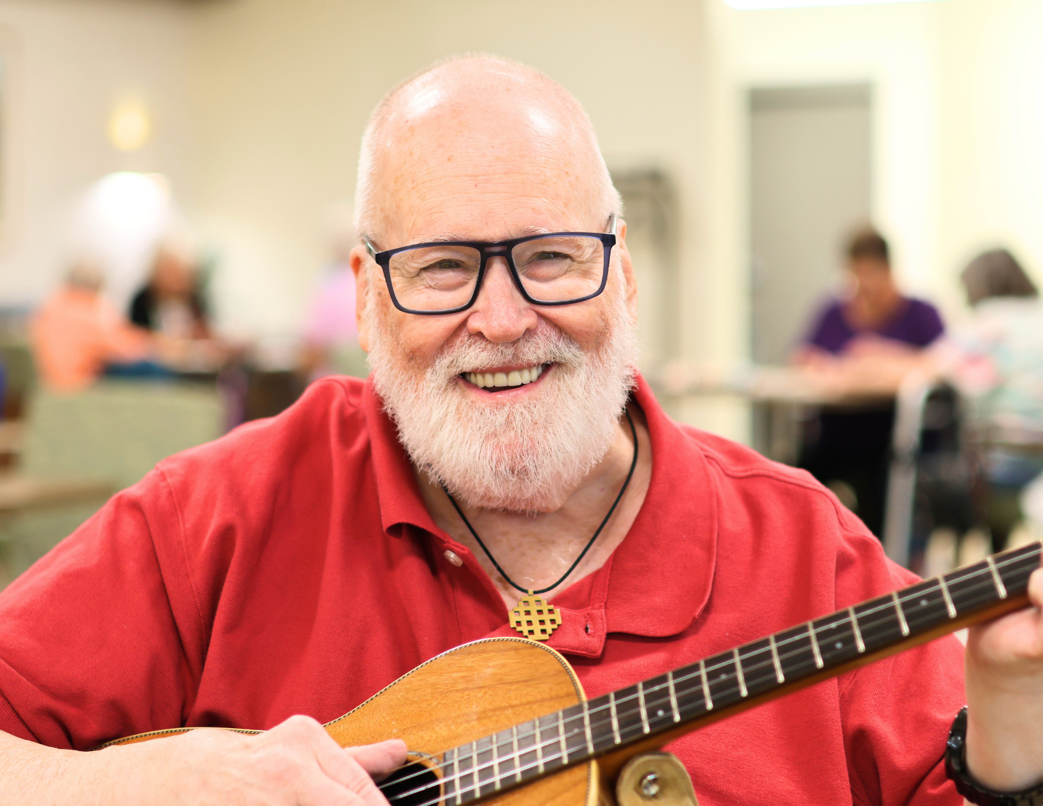 An elderly man with glasses, a white beard, and a red shirt smiling while playing a guitar in Vintage Housing with other people in the background.