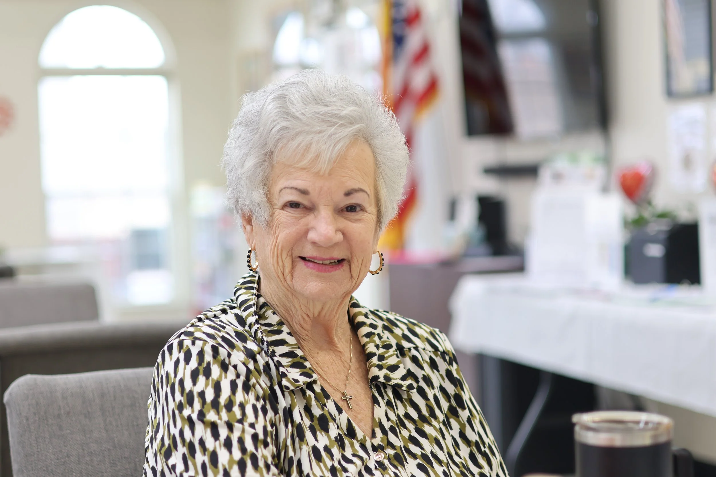 An elderly woman with short gray hair, wearing a patterned blouse sitting in the social room of Vintage Housing.
