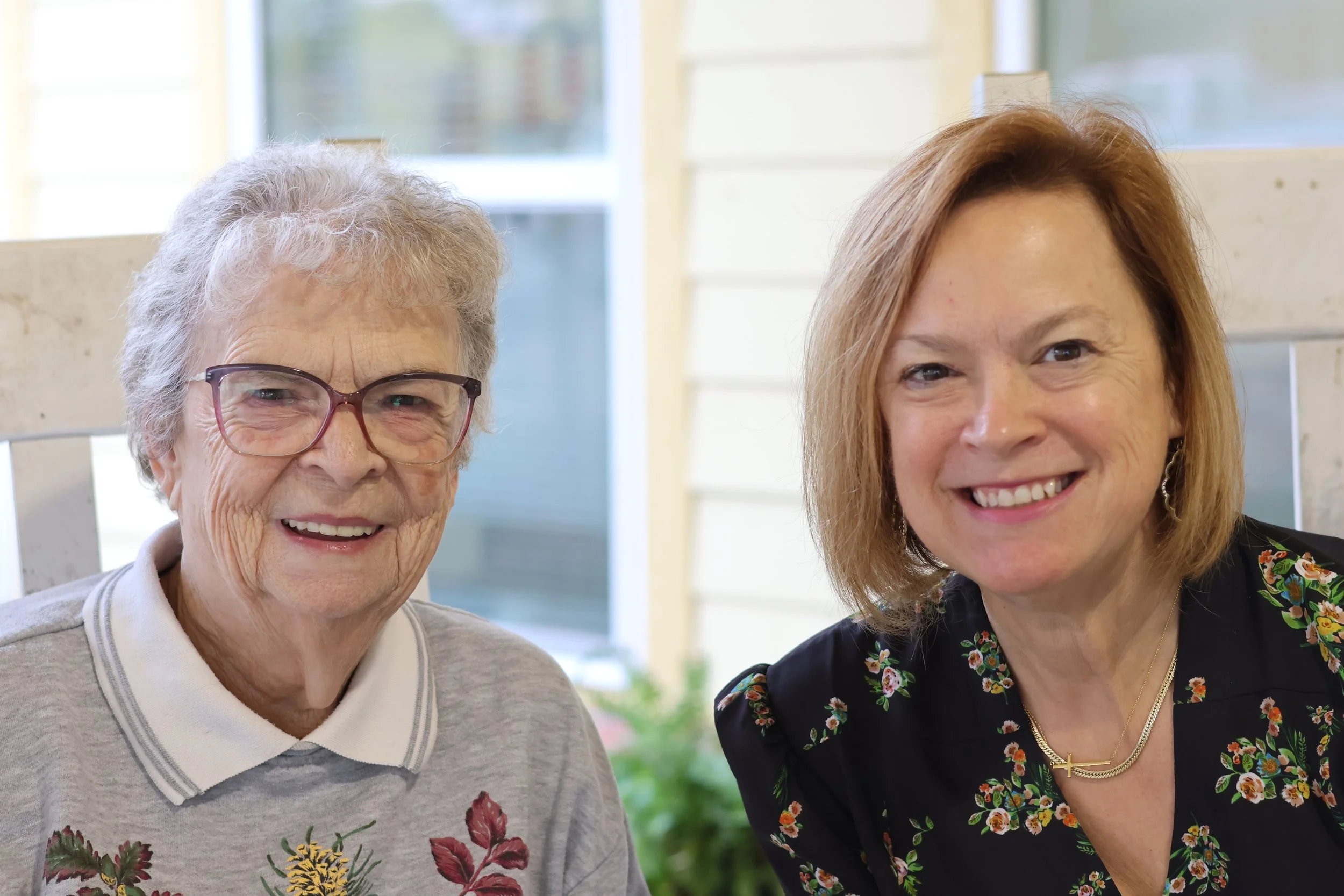 Two women smiling, one elderly woman with glasses and curly gray hair wearing a light gray sweater with floral embroidery, and one middle-aged woman with shoulder-length blonde hair wearing a black floral blouse and gold necklace. They are sitting outdoors near a house with a window.