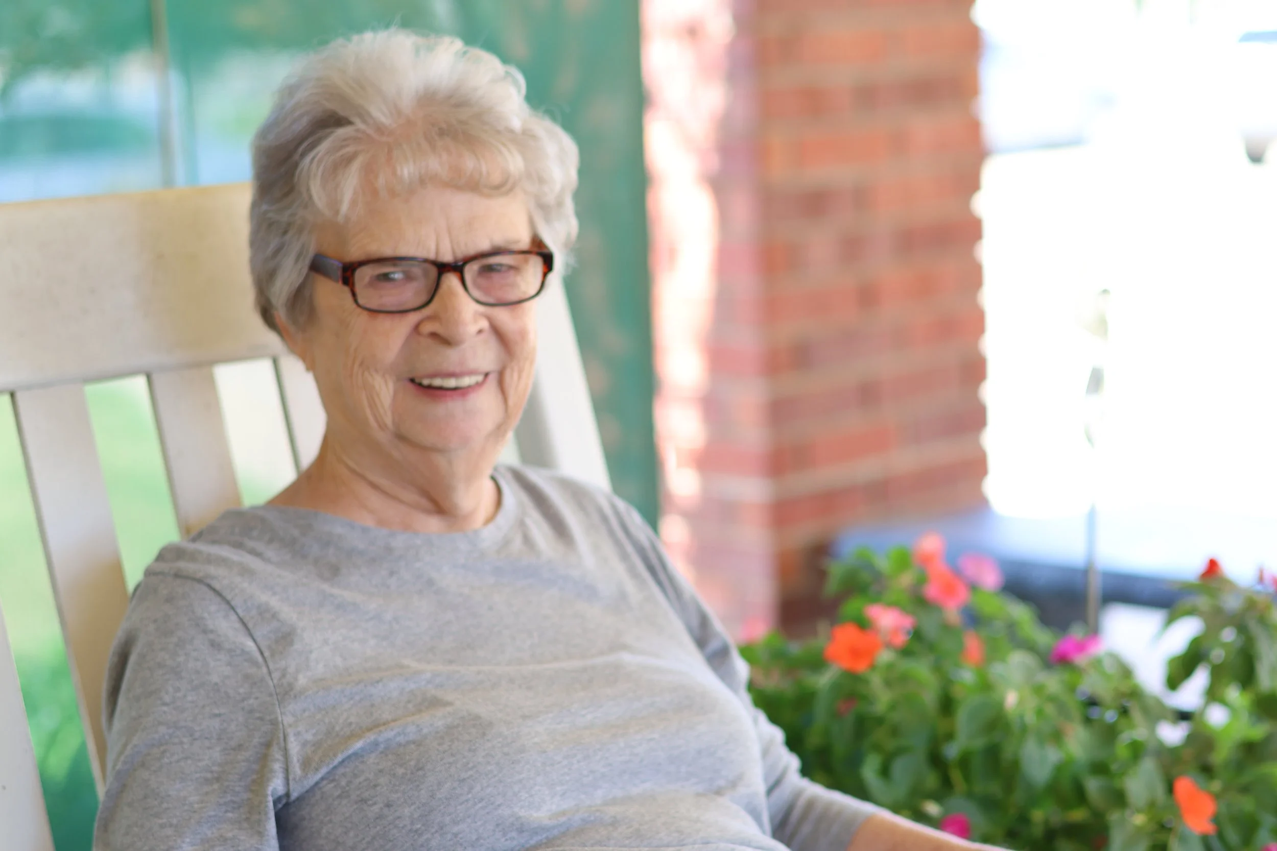 An elderly woman with glasses sitting on a porch, smiling, with potted pink and orange flowers nearby and a brick wall in the background.