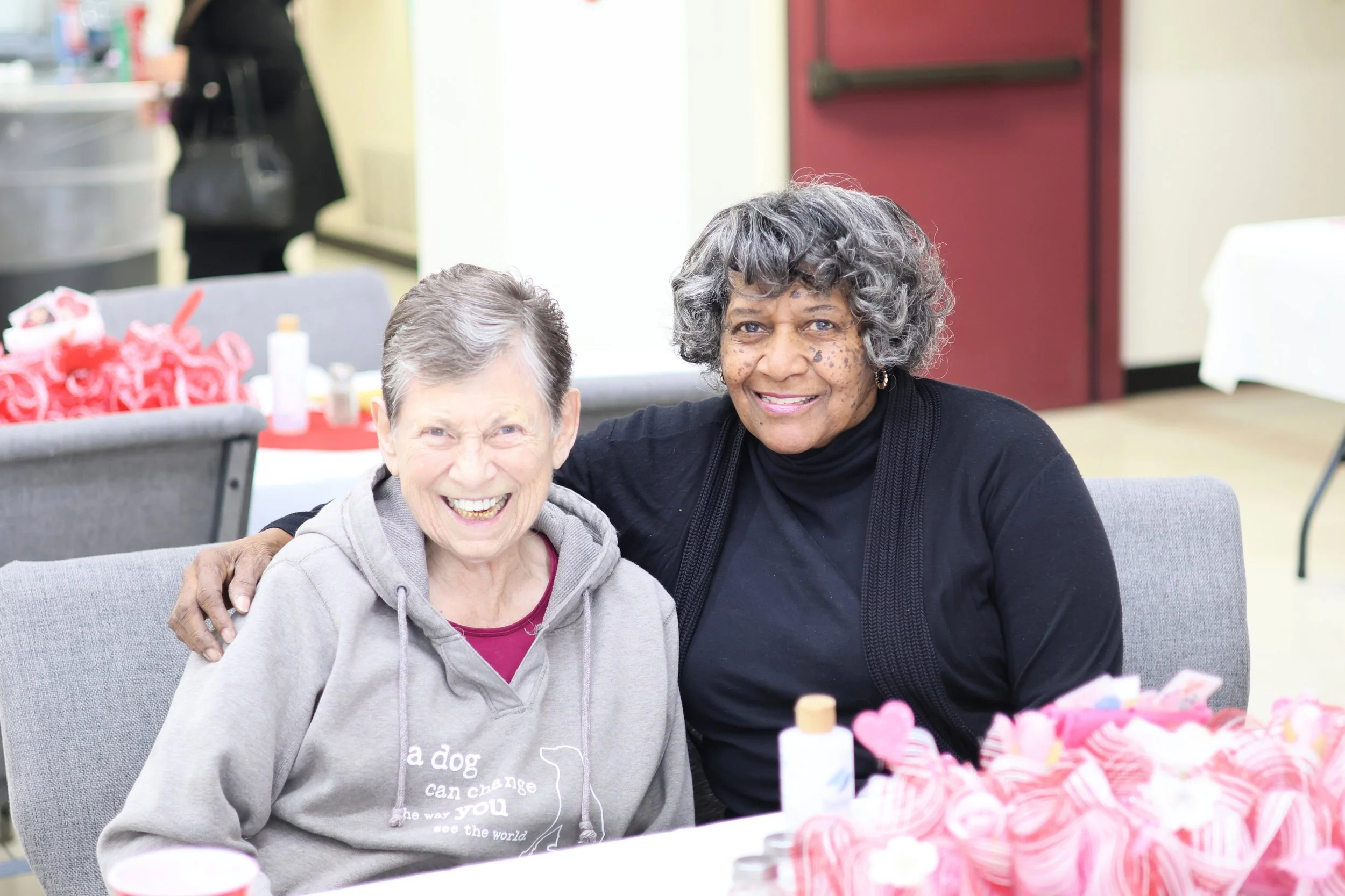 Two smiling women sitting together at a table, one elderly with gray hair and the other middle-aged with curly gray hair, in an indoor setting, possibly at a social gathering or event.