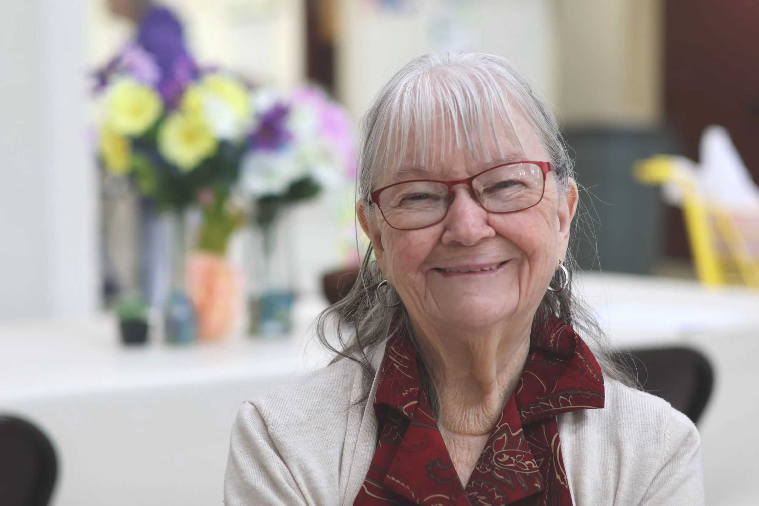 A smiling elderly woman with gray hair, glasses, and earrings, sitting indoors with a blurred background of flowers and furniture.