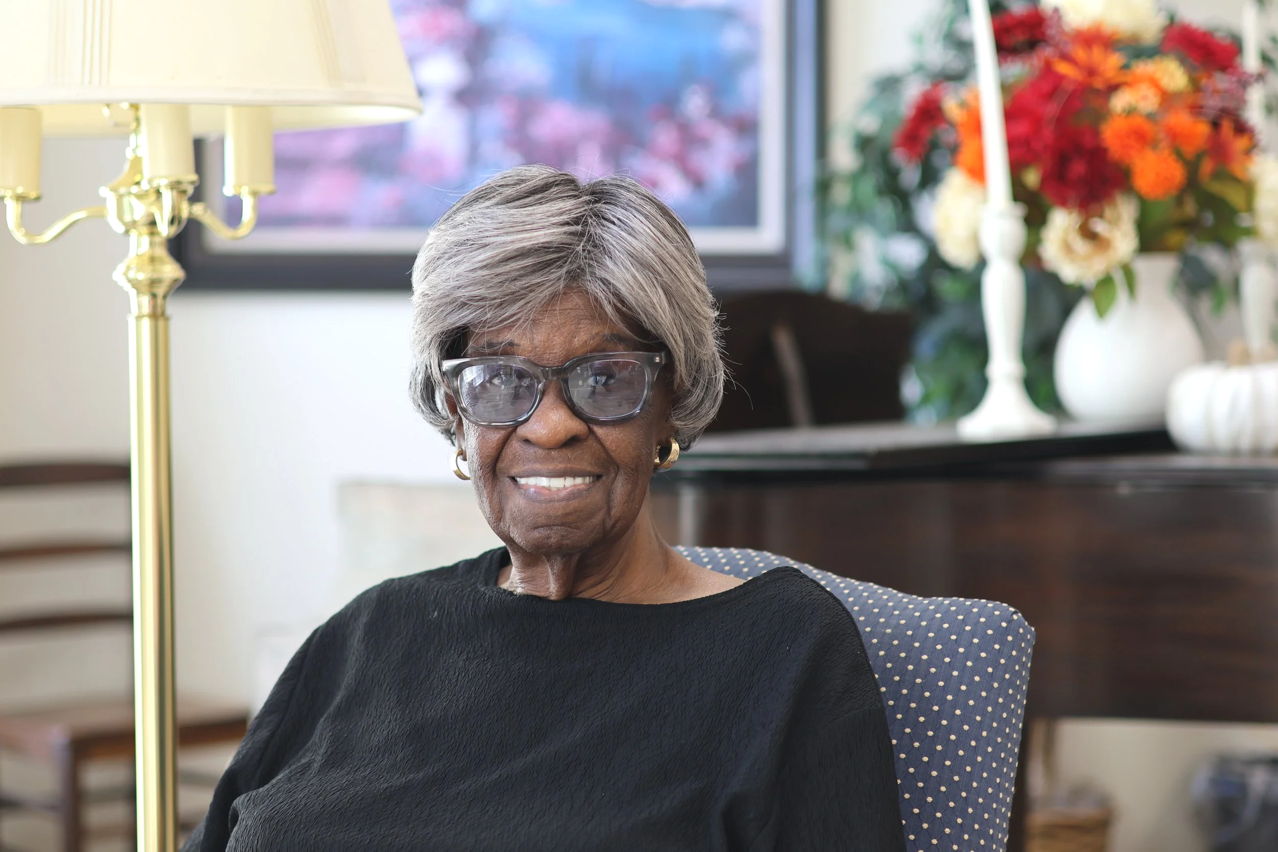 An elderly woman with gray hair, glasses, and gold earrings, smiling while sitting in a living room with a lamp and colorful floral arrangement in the background.