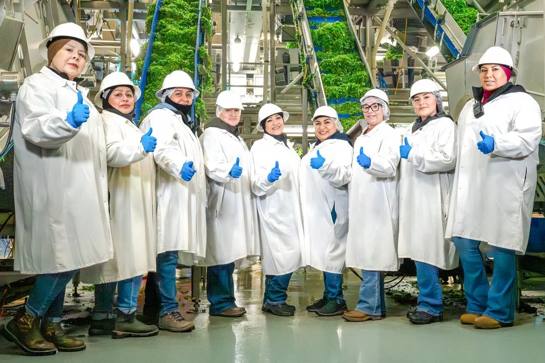 Group of women factory workers in white coats, hard hats, and blue gloves inside an industrial plant, giving thumbs up pose.