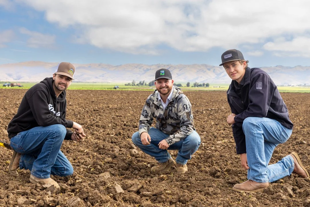 Three young men crouching in a plowed field with mountains and cloudy sky in the background.