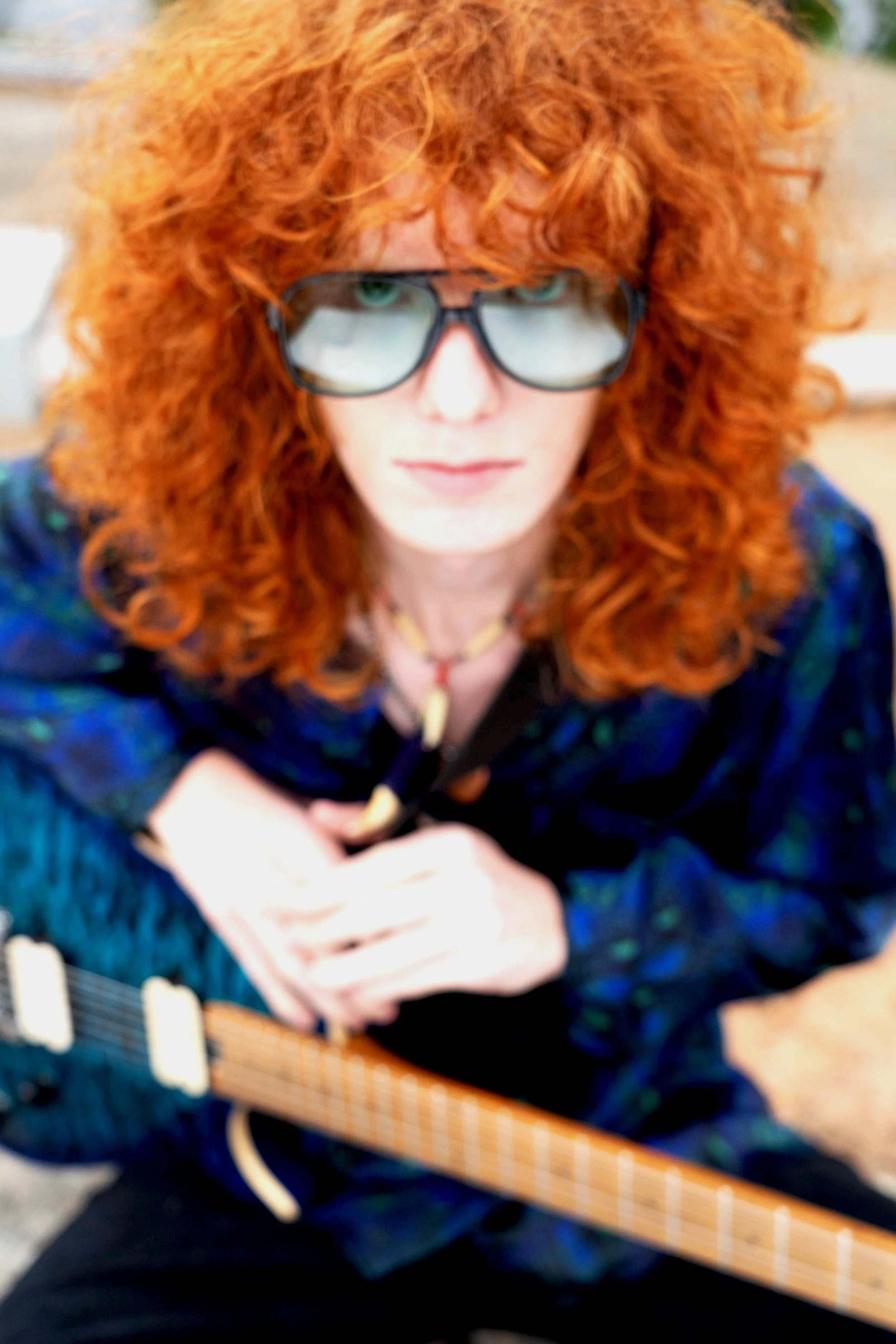 Person with long curly red hair wearing large glasses, holding a guitar, looking up at the camera.