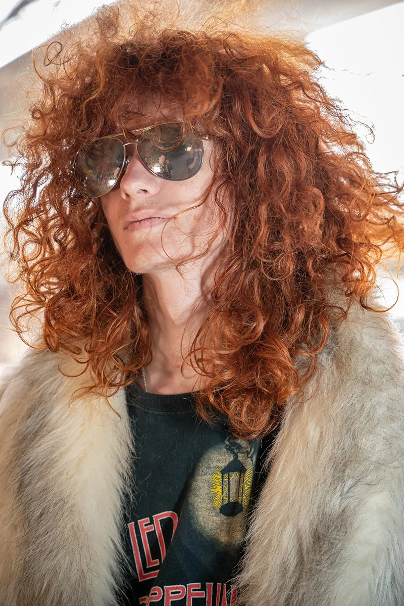 A person with curly red hair wearing sunglasses, a fur coat, and a black t-shirt with a graphic design.
