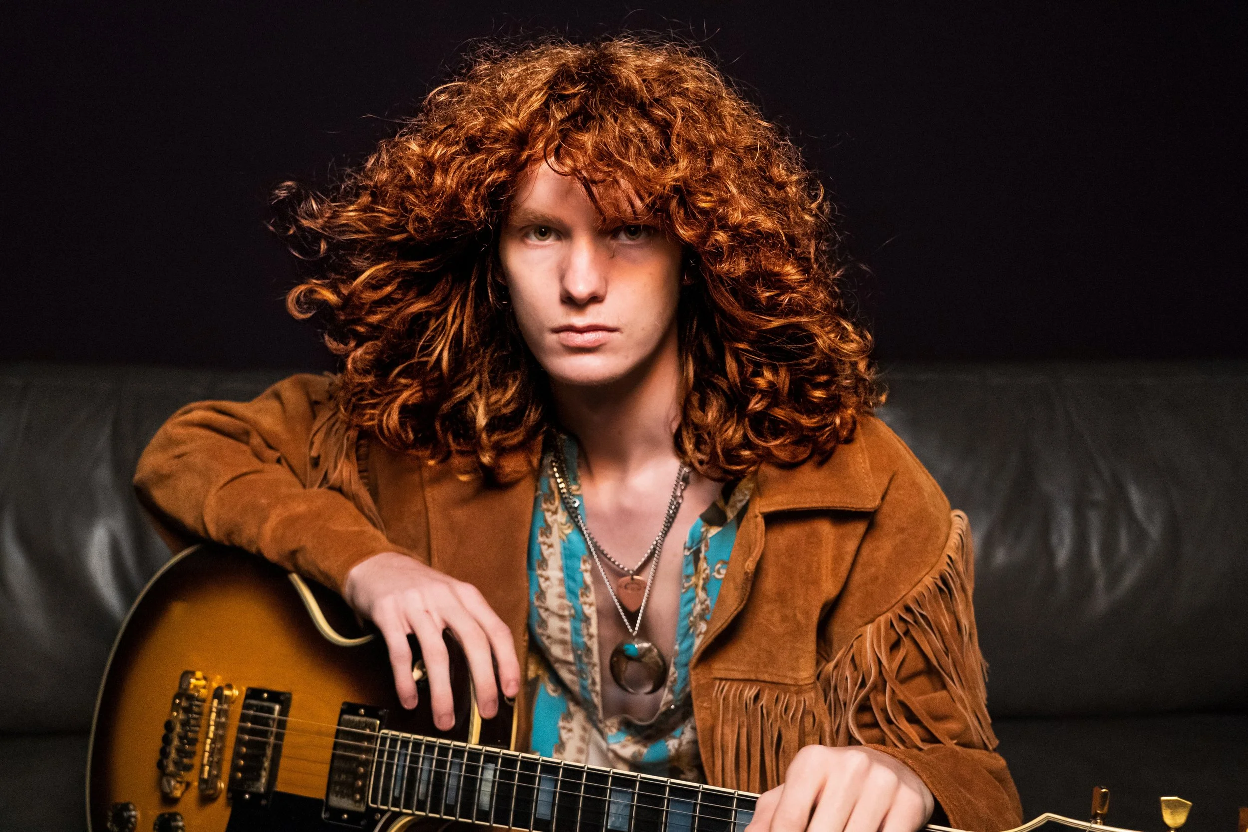 A woman with curly red hair and a serious expression, holding a guitar, sitting on a black leather couch against a dark background.